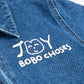 A close-up of the white "JOY BOBO CHOSES" logo printed on the left chest of a dark blue denim jumpsuit. The playful design features a hand-drawn smiling face alongside the text. The denim fabric has visible stitching details and a slightly worn texture, with part of the structured collar showing at the top of the frame.