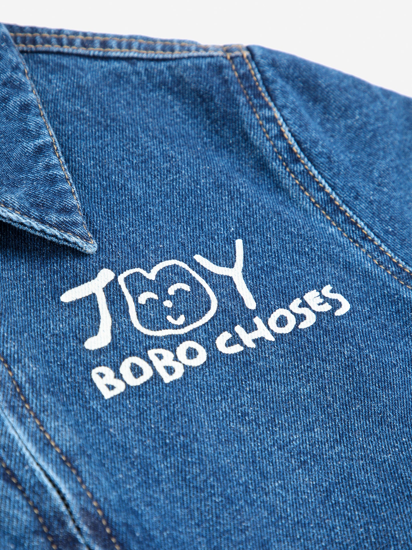 A close-up of the white "JOY BOBO CHOSES" logo printed on the left chest of a dark blue denim jumpsuit. The playful design features a hand-drawn smiling face alongside the text. The denim fabric has visible stitching details and a slightly worn texture, with part of the structured collar showing at the top of the frame.