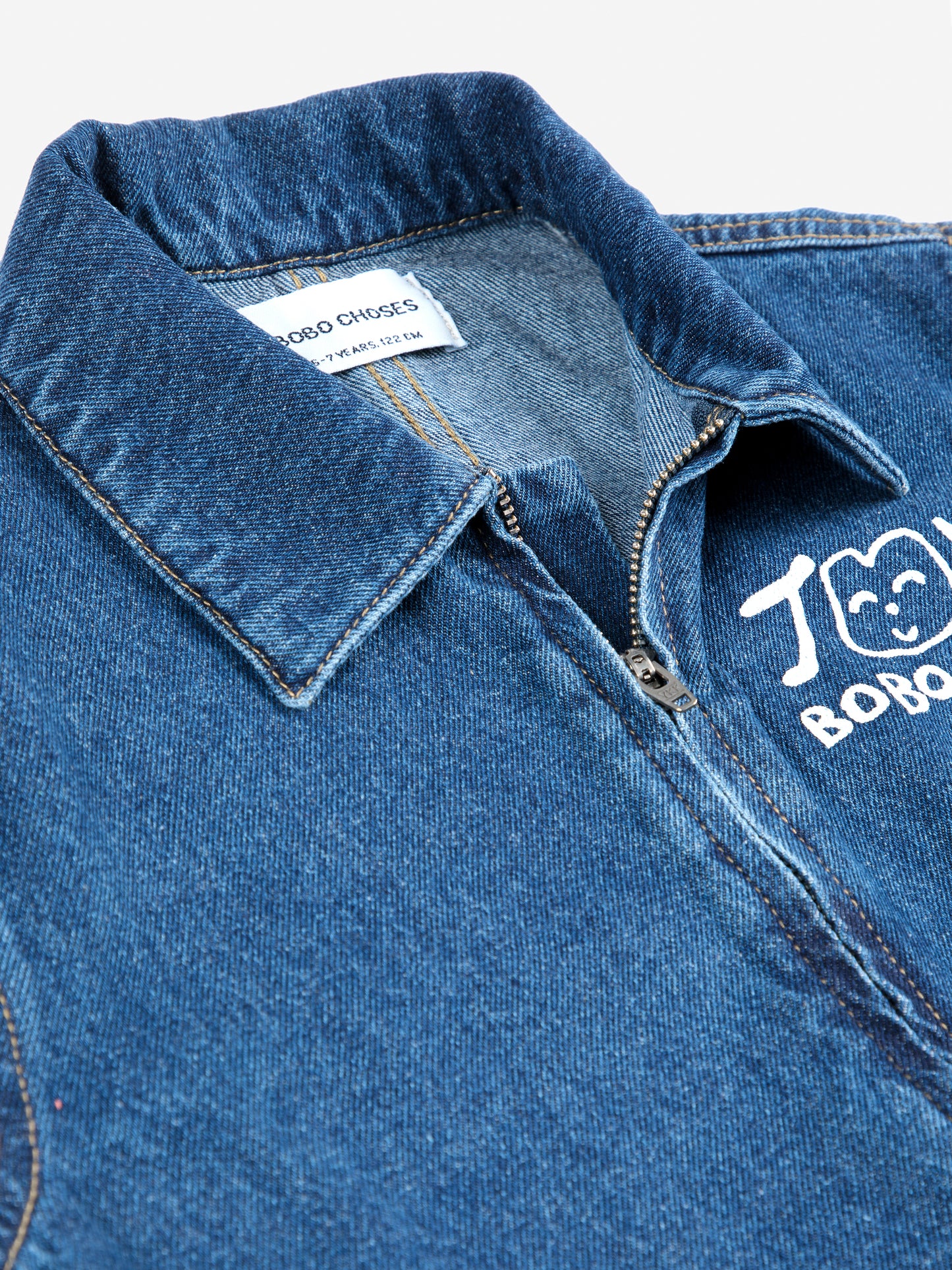 A close-up of the collar and upper chest area of a dark blue denim jumpsuit. The structured collar has visible stitching, and a partially unzipped metal zipper runs down the front. A white "JOY BOBO CHOSES" logo with a smiling face is printed on the left chest. Inside the collar, a white label displays the brand name "BOBO CHOSES" and size details. The fabric has a slightly worn texture.