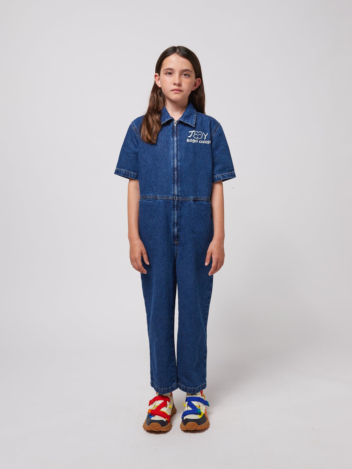 A young child with long dark hair stands facing forward, wearing a dark blue denim jumpsuit with short sleeves, a structured collar, and a front zip closure. The "JOY BOBO CHOSES" logo with a smiling face is printed in white on the left chest. The jumpsuit has a relaxed fit with visible stitching details. They are also wearing chunky sandals with multicoloured straps in red, blue, and yellow, and thick brown soles. The background is plain white