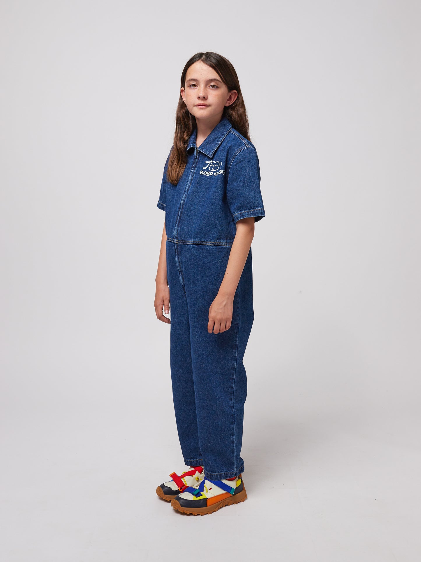 A young child with long dark hair stands slightly turned to the side, wearing a dark blue denim jumpsuit with short sleeves, a structured collar, and a front zip closure. The "JOY BOBO CHOSES" logo with a smiling face is printed in white on the left chest. The jumpsuit has a relaxed fit with visible stitching details. They are also wearing chunky sandals with multicoloured straps in red, blue, and yellow, and thick brown soles. The background is plain white.