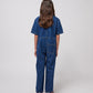 A young child with long dark hair is seen from the back, wearing a dark blue denim jumpsuit with short sleeves and a relaxed fit. The jumpsuit features visible stitching details, a structured collar, and a single square patch pocket on the right side of the lower back. They are also wearing chunky sandals with multicoloured straps and thick brown soles. The background is plain white.