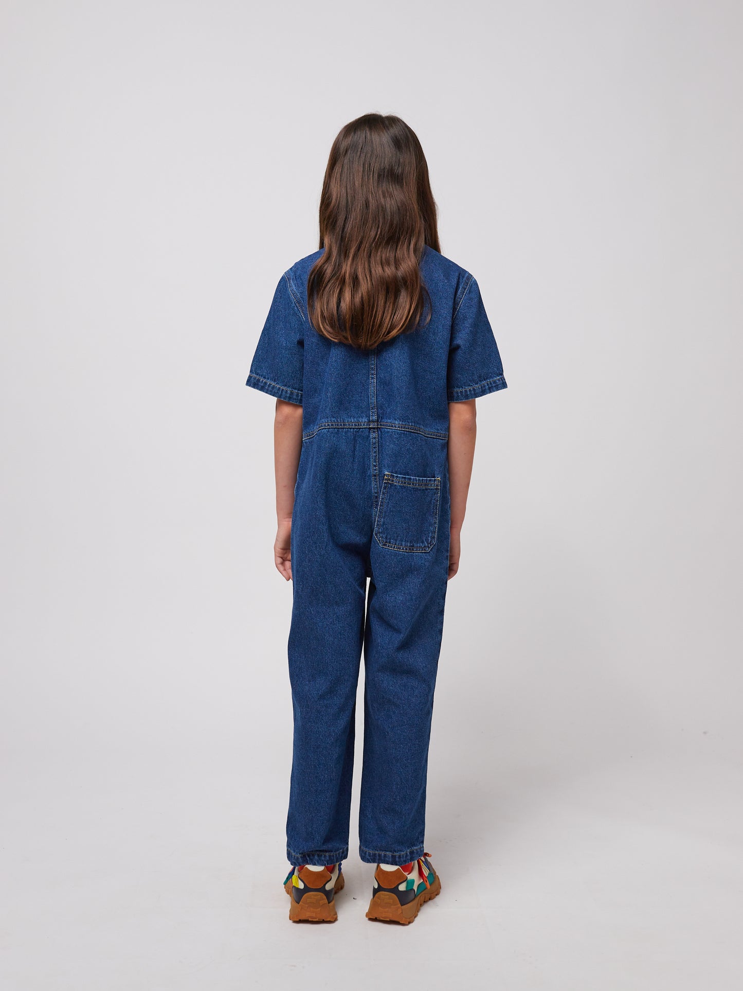 A young child with long dark hair is seen from the back, wearing a dark blue denim jumpsuit with short sleeves and a relaxed fit. The jumpsuit features visible stitching details, a structured collar, and a single square patch pocket on the right side of the lower back. They are also wearing chunky sandals with multicoloured straps and thick brown soles. The background is plain white.