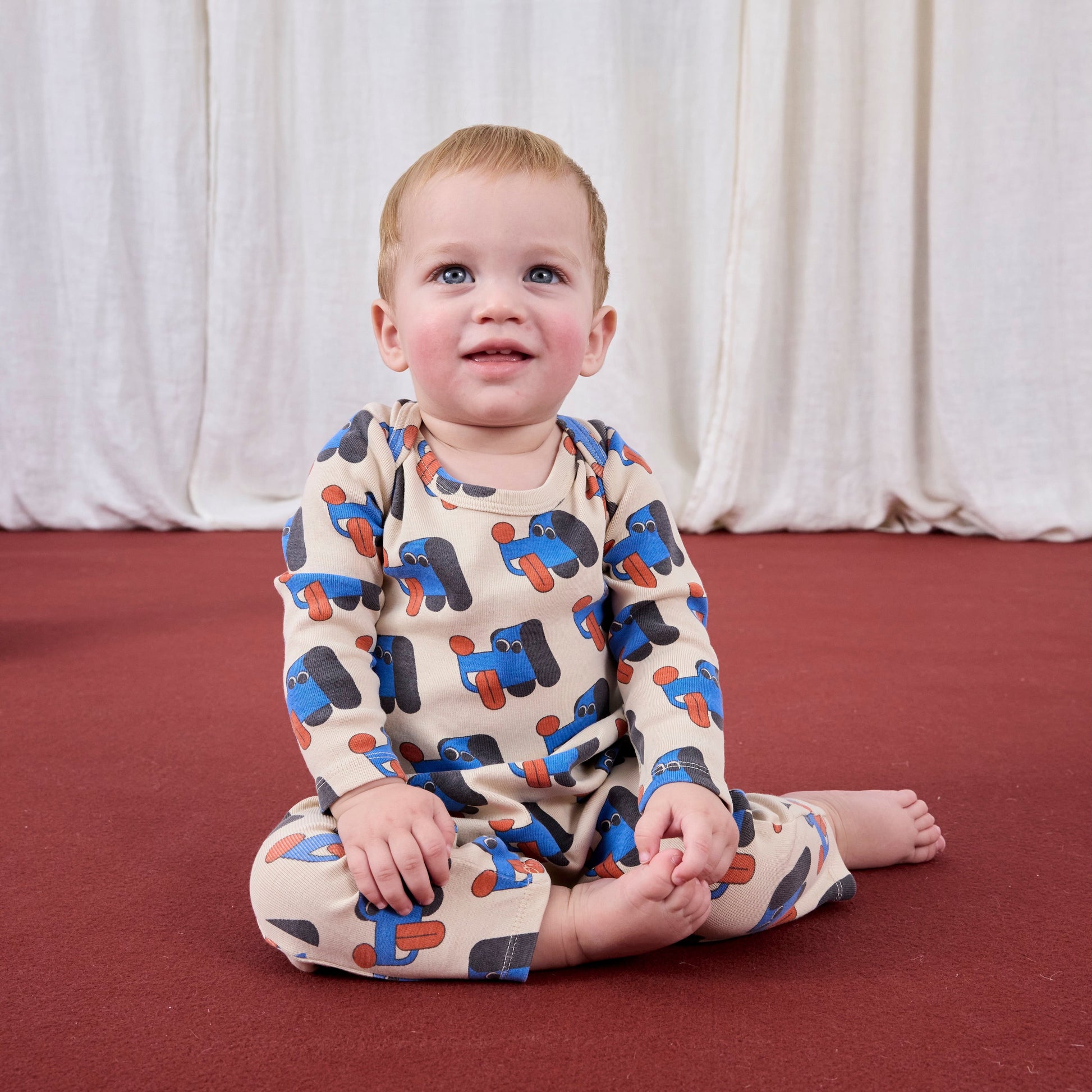 Baby sitting on a red floor wearing a patterned outfit with a white curtain background