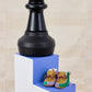 Large black chess piece on a blue platform with baby shoes below, against a beige wall.