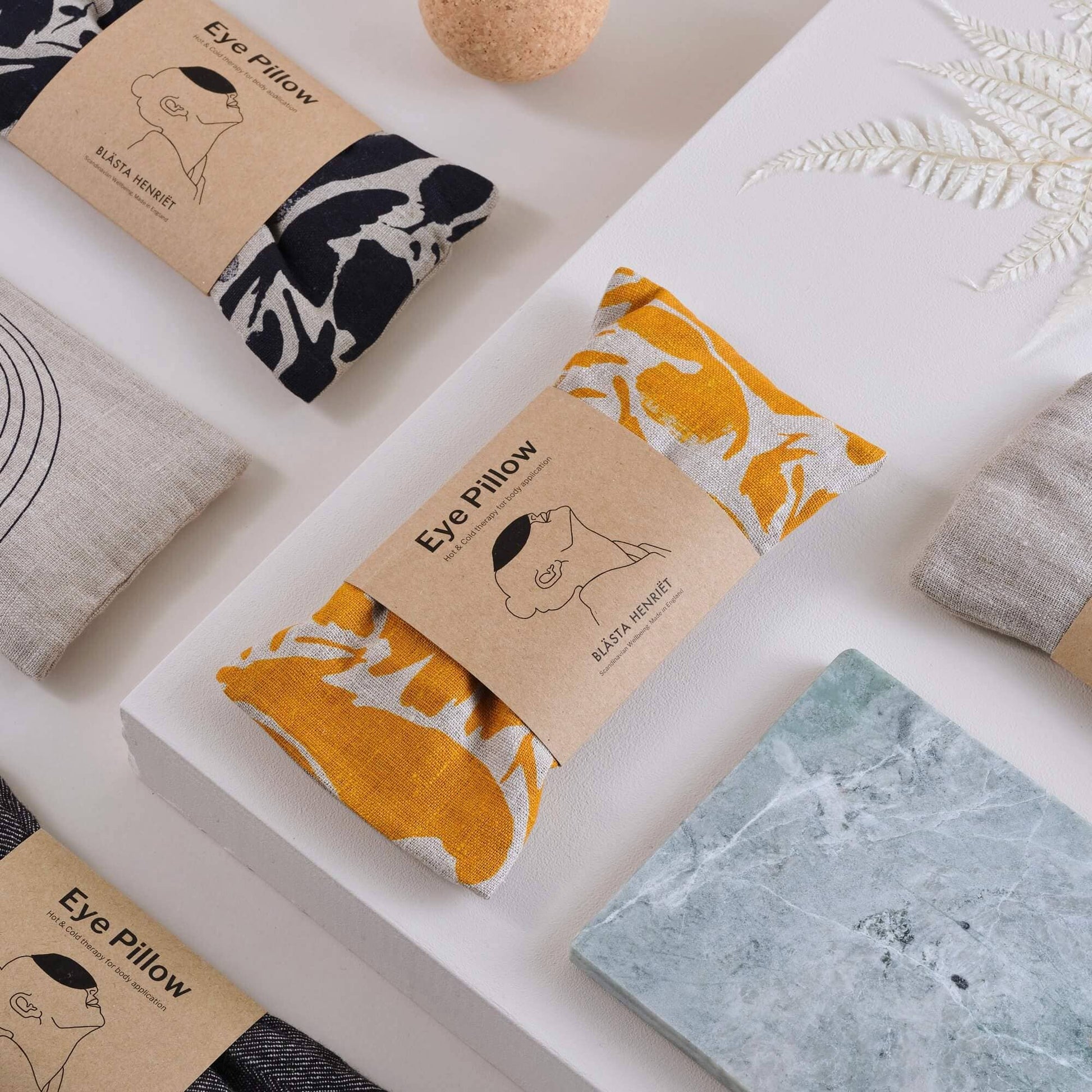 Flat lay of Blästa Henriët eye pillows, featuring both navy and yellow abstract prints, arranged with other wellness items on a minimalist white surface.