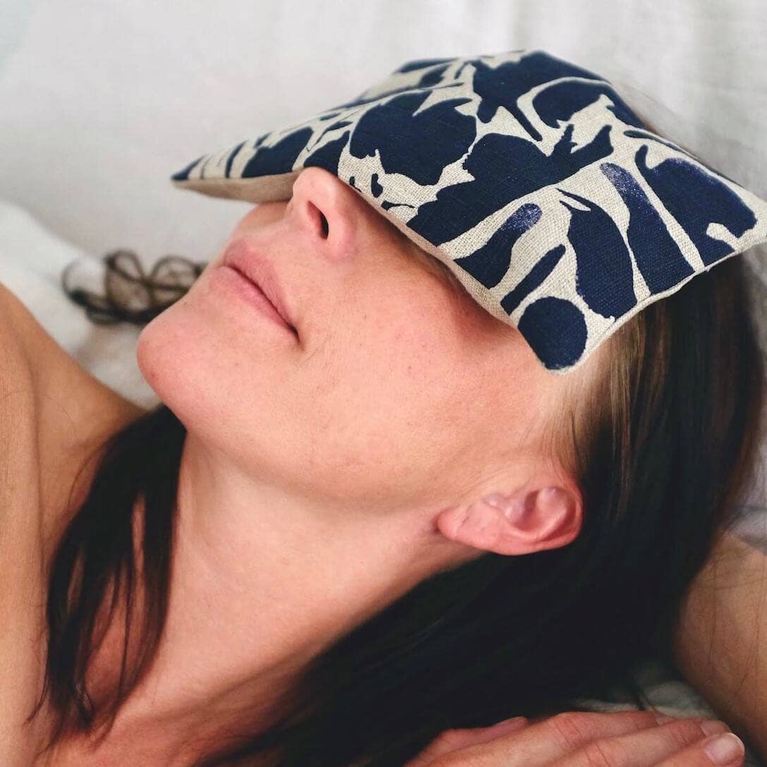 Person reclining with a navy and white abstract-patterned linen eye pillow placed over their eyes, promoting rest and relaxation.