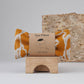Blästa Henriët eye pillow with a bold yellow and white abstract print, displayed in eco-friendly packaging on a wooden stand with a natural birch backdrop.