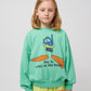 A young child with blonde hair wears the green sweatshirt, paired with textured yellow trousers. The sweatshirt has a relaxed fit with balloon sleeves.