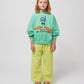 The child stands facing the camera, wearing the sweatshirt with wide-leg yellow checkered trousers and red sneakers.