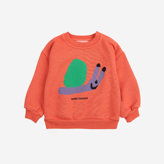 A Bobo Choses baby sweatshirt in coral, featuring a cheerful snail illustration with a green shell and a purple body with a smile. Below the snail, "BOBO CHOSES" is printed in black. The sweatshirt has ribbed cuffs and a relaxed fit.