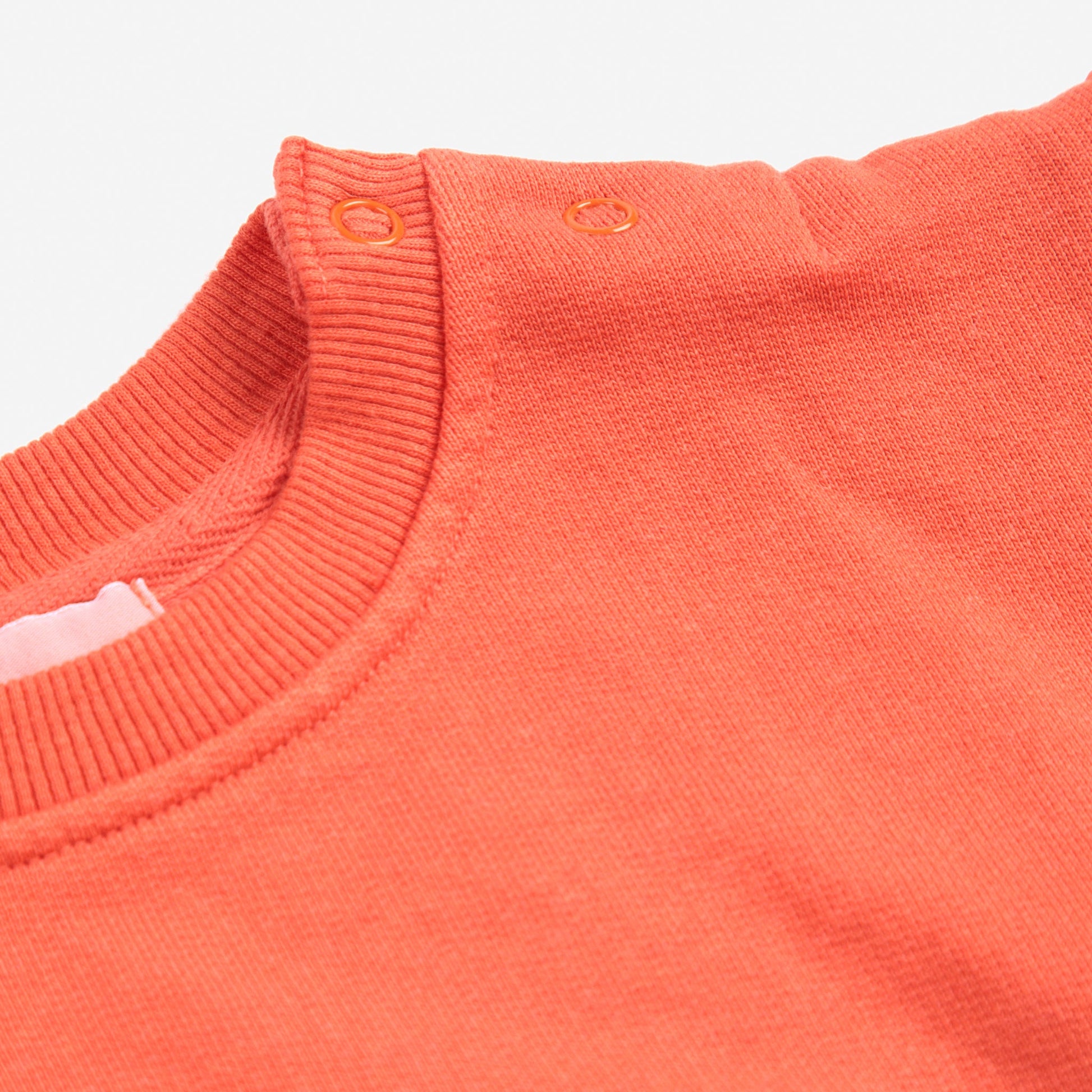 A close-up of the neckline of a coral Bobo Choses baby sweatshirt, featuring ribbed trim and two small snap buttons on the shoulder for easy dressing. Part of a playful snail print is visible on the fabric.