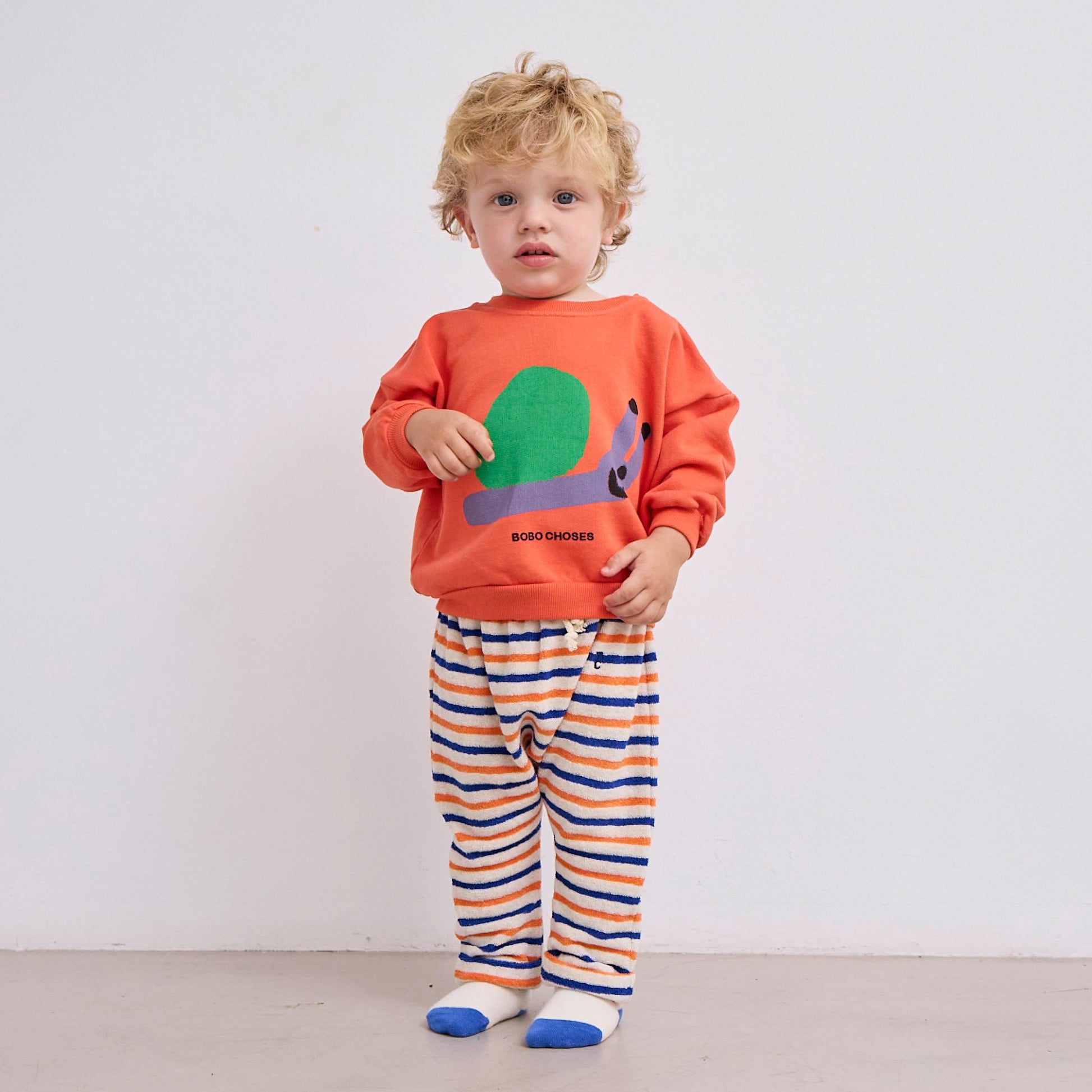 A toddler wearing the Bobo Choses snail sweatshirt, paired with striped terry cloth trousers in orange, blue, and cream. The relaxed fit of the sweatshirt and soft texture of the trousers create a cosy outfit.