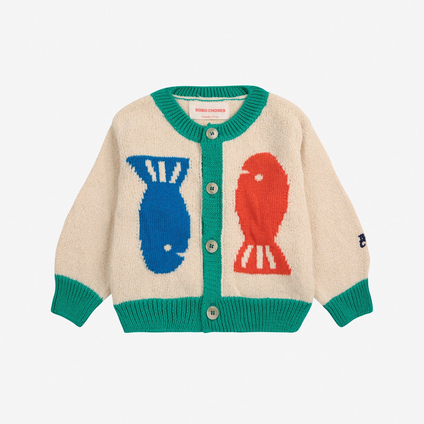 A baby cardigan in natural beige with a playful fish motif on the front, featuring a blue fish on one side and a red fish on the other. It has green ribbed trim at the neckline, cuffs, and hem, with a button-down front.