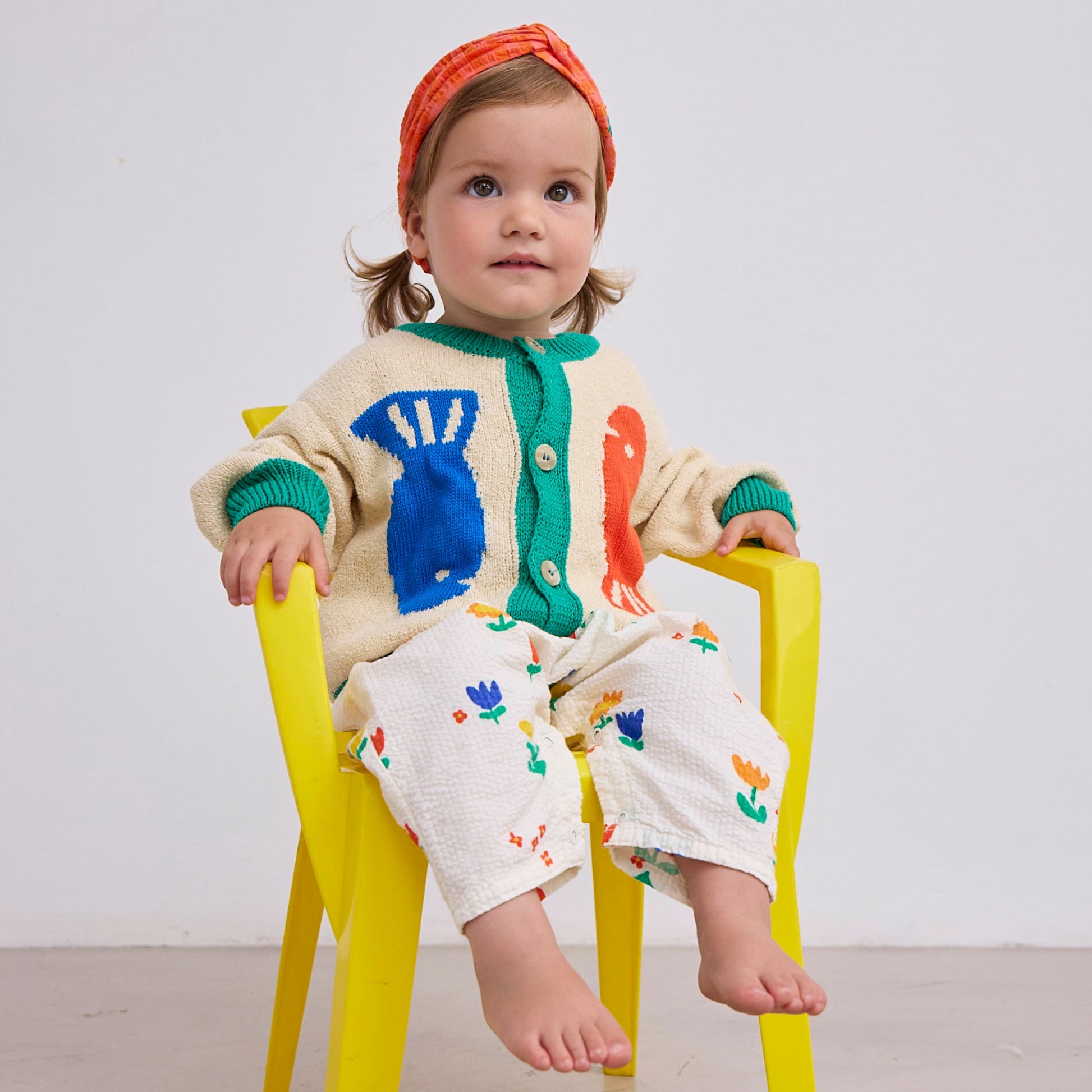 A toddler sits on a yellow chair, wearing the cardigan with its playful fish motif and green trim. The child pairs it with patterned trousers and an orange headband.