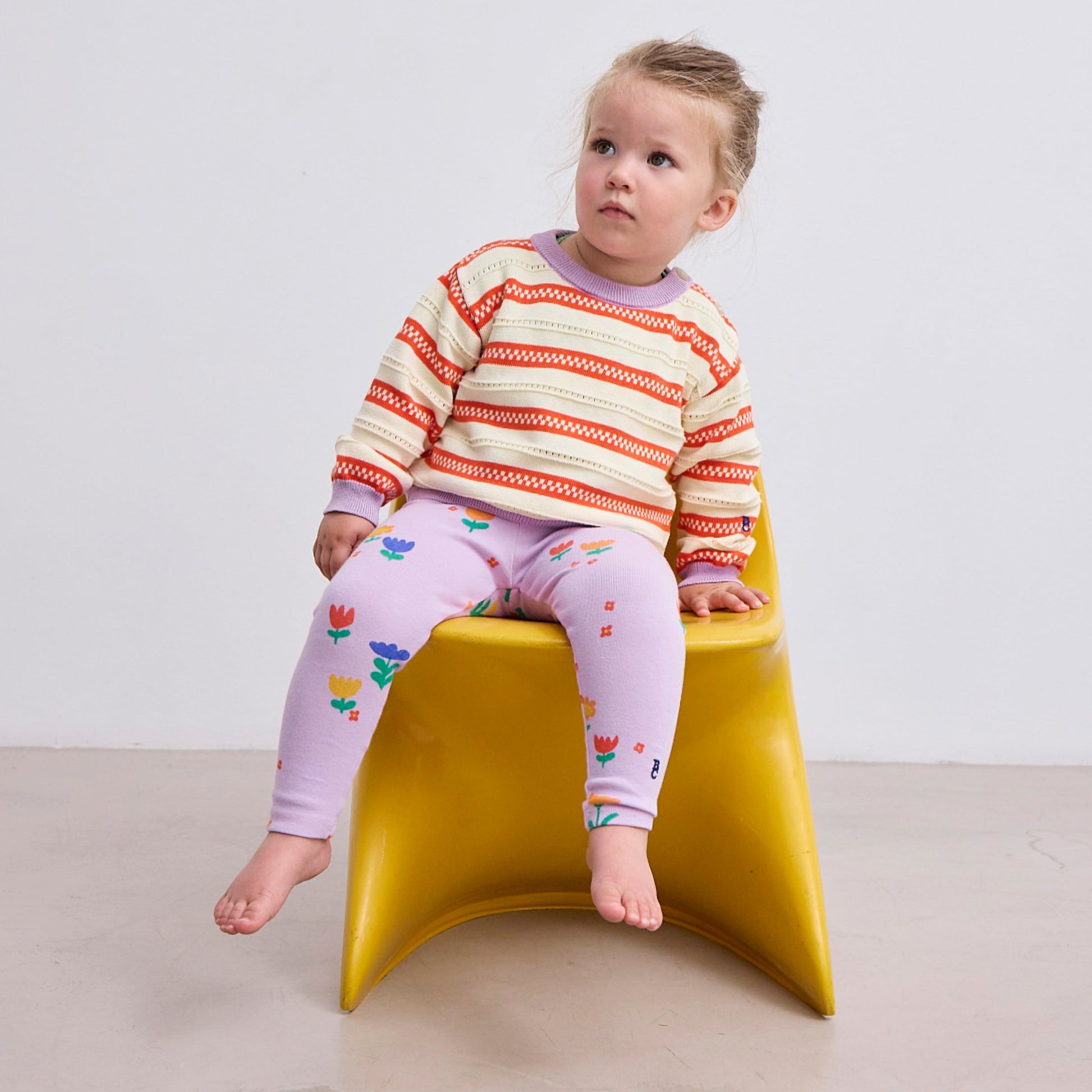 A young child sits on a yellow chair, wearing the striped jumper paired with lilac leggings featuring a floral print.