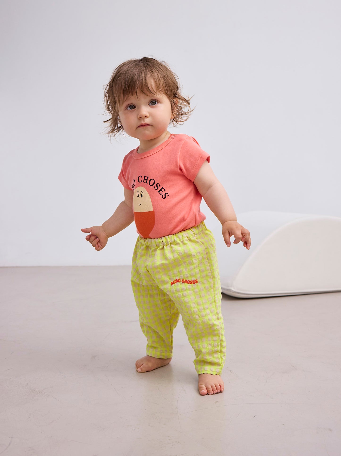 A toddler with short, wavy brown hair wears the gingham baby pants along with a coral pink T-shirt featuring a smiling face graphic and the "BOBO CHOSES" logo. The child is standing barefoot on a neutral floor.