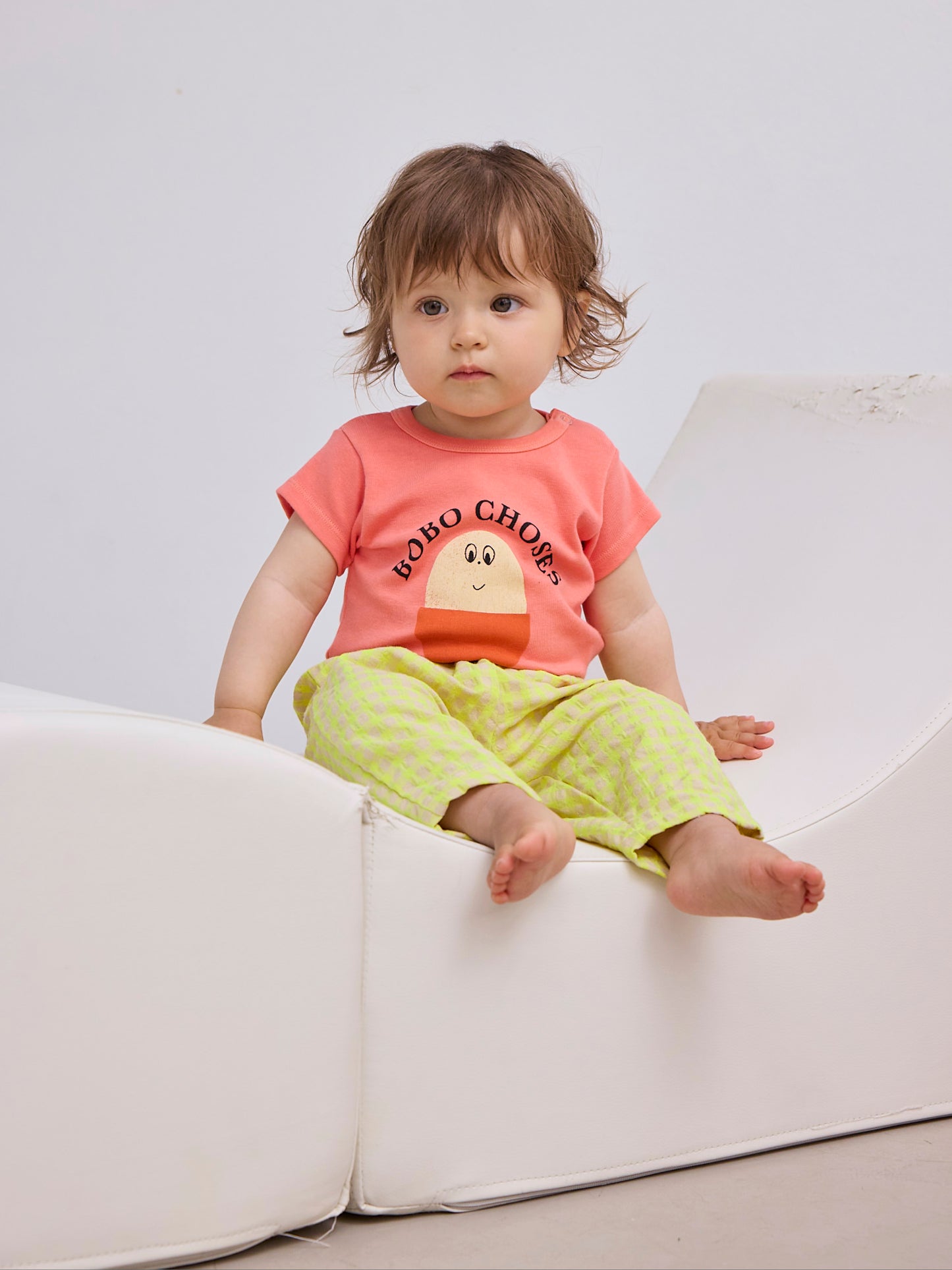 The same toddler sits on a white, modern-shaped cushioned seat while wearing the yellow gingham pants and coral T-shirt. Their legs are stretched out and slightly bent.