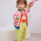 The toddler is now wearing a pink knit cardigan with blue and green trim over the coral T-shirt, paired with the gingham pants. They are standing while holding onto an adult’s hand.