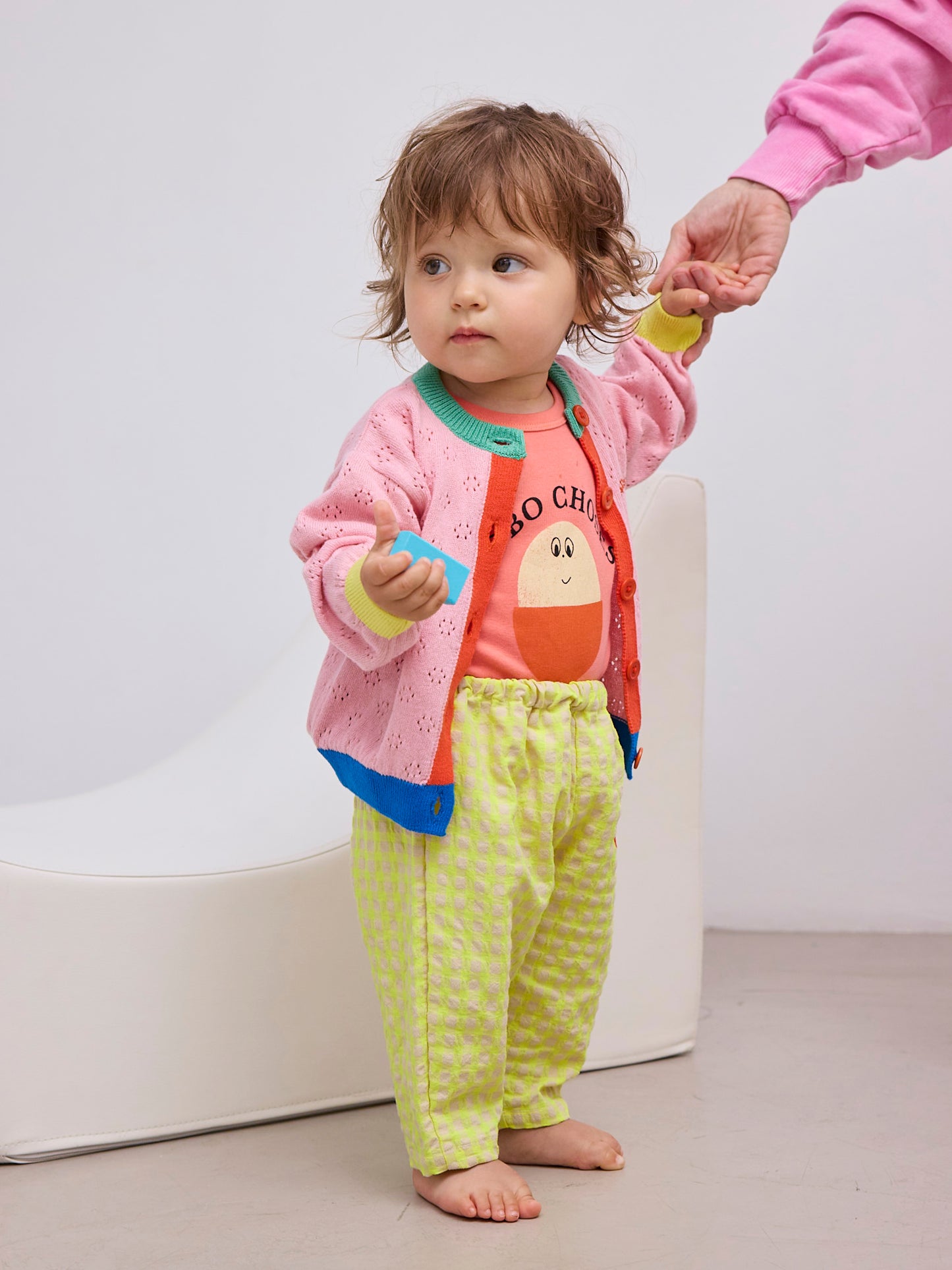 The toddler is now wearing a pink knit cardigan with blue and green trim over the coral T-shirt, paired with the gingham pants. They are standing while holding onto an adult’s hand.