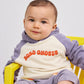 A baby sitting in a yellow chair wearing the Bobo Choses terry cloth hoodie. The relaxed fit and plush texture of the hoodie are visible, with the baby also wearing matching soft purple trousers.