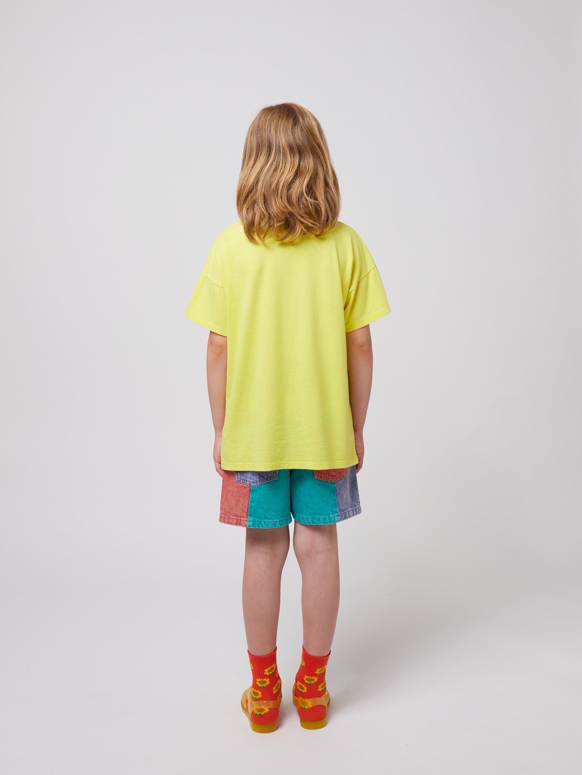 A young girl with shoulder-length blonde hair wearing the yellow T-shirt with multicolour shorts and bright sandals.
Bobo Choses Funny Snail T-Shirt (Back View) – The same girl from behind, showing the T-shirt’s fit and length.