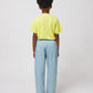  A child standing with their back to the camera against a plain white background, wearing a bright yellow Bobo Choses Funny Snail T-Shirt and relaxed-fit light blue trousers with two back pockets. The child's curly hair is visible above the collar of the shirt, and they are wearing red canvas sneakers with white soles.