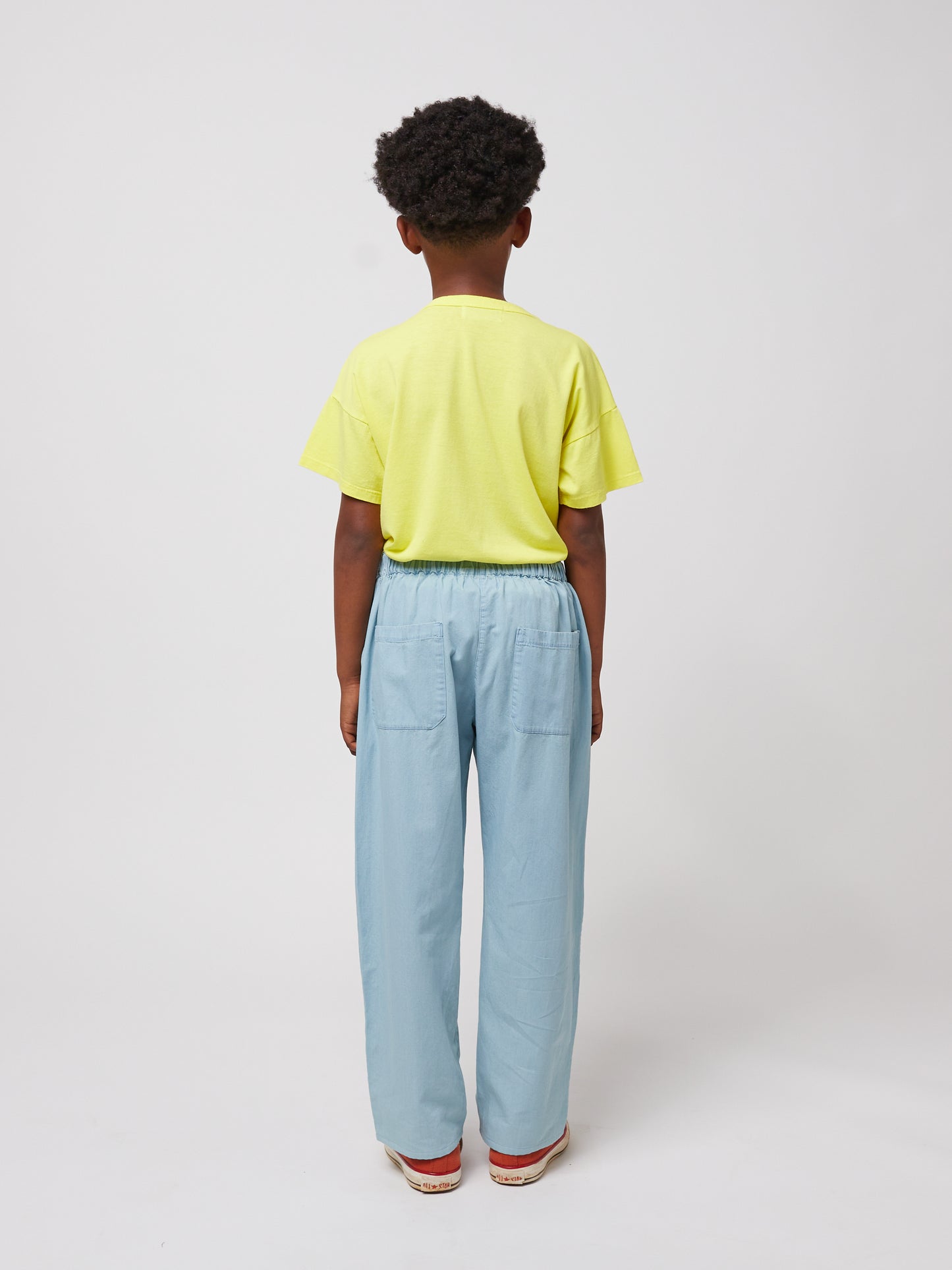  A child standing with their back to the camera against a plain white background, wearing a bright yellow Bobo Choses Funny Snail T-Shirt and relaxed-fit light blue trousers with two back pockets. The child's curly hair is visible above the collar of the shirt, and they are wearing red canvas sneakers with white soles.