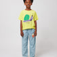A child standing against a plain background, wearing the Bobo Choses Funny Snail T-Shirt in yellow with a playful green and purple snail print on the front. They are also wearing light blue trousers featuring a small illustrated face design on one leg and red canvas sneakers with white laces. The child has a bright, happy expression.