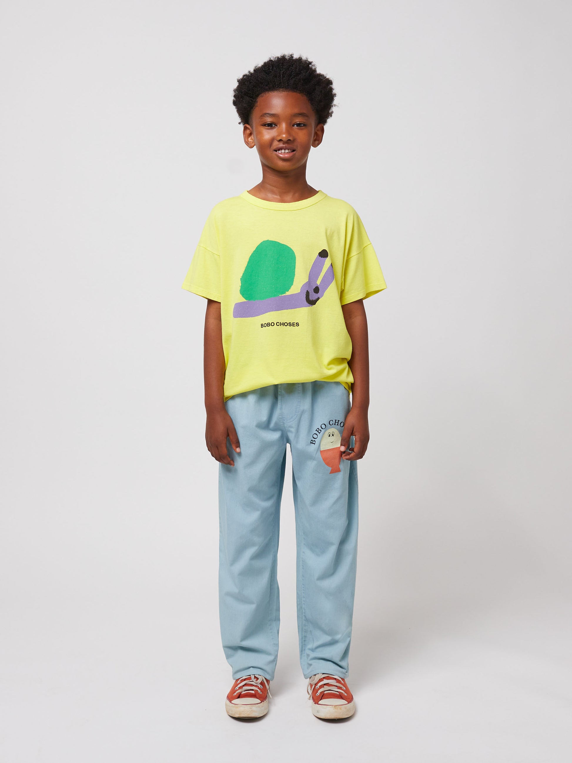 A child standing against a plain background, wearing the Bobo Choses Funny Snail T-Shirt in yellow with a playful green and purple snail print on the front. They are also wearing light blue trousers featuring a small illustrated face design on one leg and red canvas sneakers with white laces. The child has a bright, happy expression.