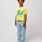 A child standing against a plain white background, wearing the Bobo Choses Funny Snail T-Shirt in bright yellow with a whimsical green and purple snail print. The shirt is paired with relaxed-fit light blue trousers featuring a small orange illustrated character on one leg. The child is also wearing red canvas sneakers with white laces and has a confident, relaxed expression.