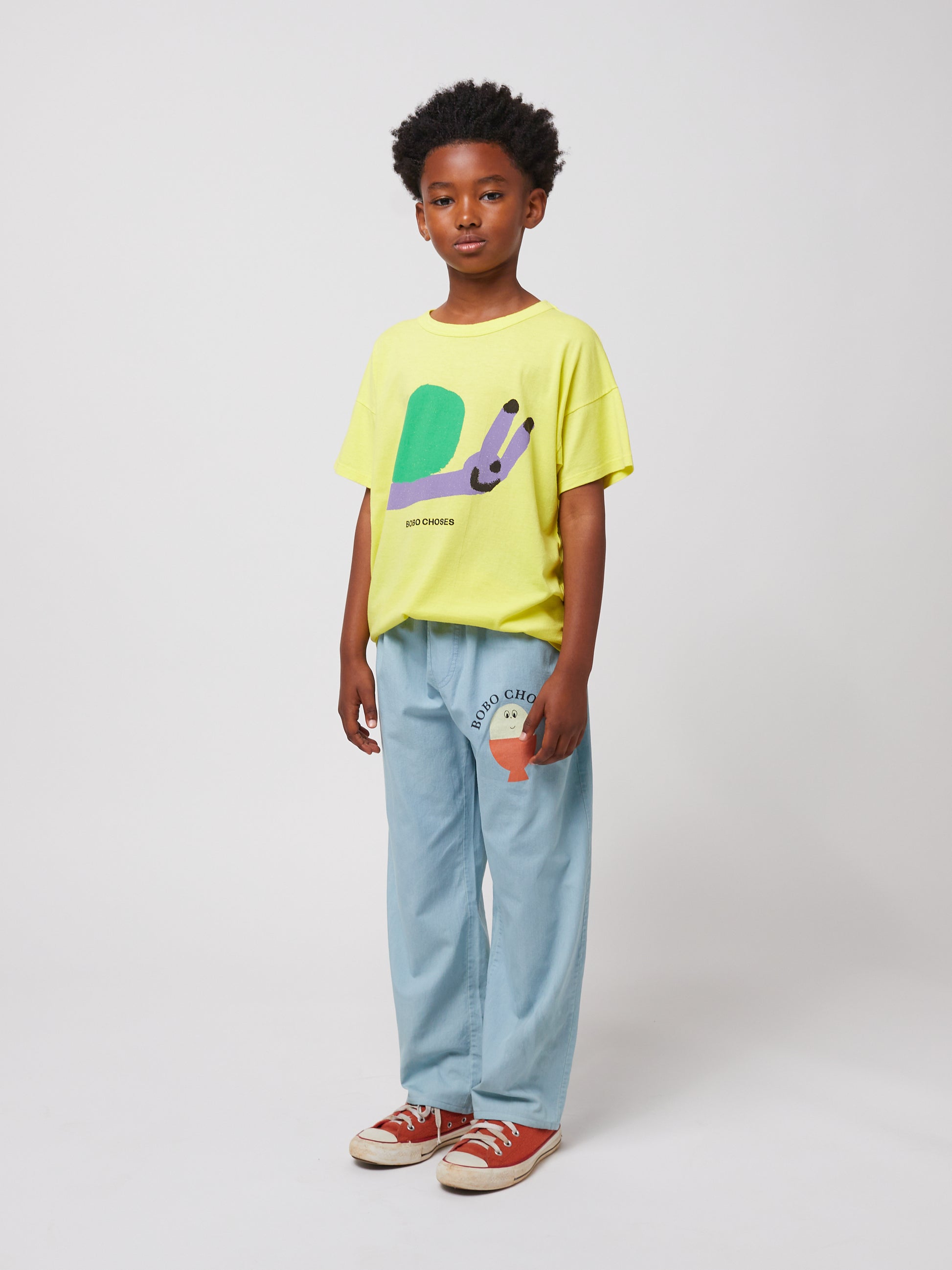 A child standing against a plain white background, wearing the Bobo Choses Funny Snail T-Shirt in bright yellow with a whimsical green and purple snail print. The shirt is paired with relaxed-fit light blue trousers featuring a small orange illustrated character on one leg. The child is also wearing red canvas sneakers with white laces and has a confident, relaxed expression.