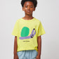 A child wearing the Bobo Choses Funny Snail T-Shirt in yellow, featuring a playful green and purple snail illustration on the front with "BOBO CHOSES" text below. The child pairs it with light blue wide-leg trousers that have a small illustrated face print on the leg.
