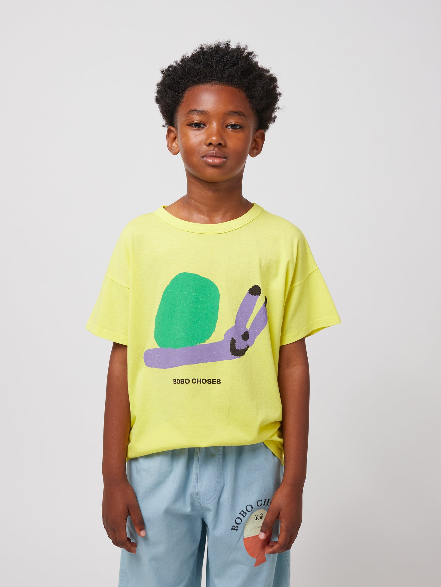 A child wearing the Bobo Choses Funny Snail T-Shirt in yellow, featuring a playful green and purple snail illustration on the front with "BOBO CHOSES" text below. The child pairs it with light blue wide-leg trousers that have a small illustrated face print on the leg.