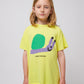 A child wearing the Bobo Choses Funny Snail T-Shirt in bright yellow. The T-shirt features a playful illustration of a smiling snail with a green shell and a lavender body, along with "BOBO CHOSES" printed in black below the design. The child is also wearing multicoloured shorts.