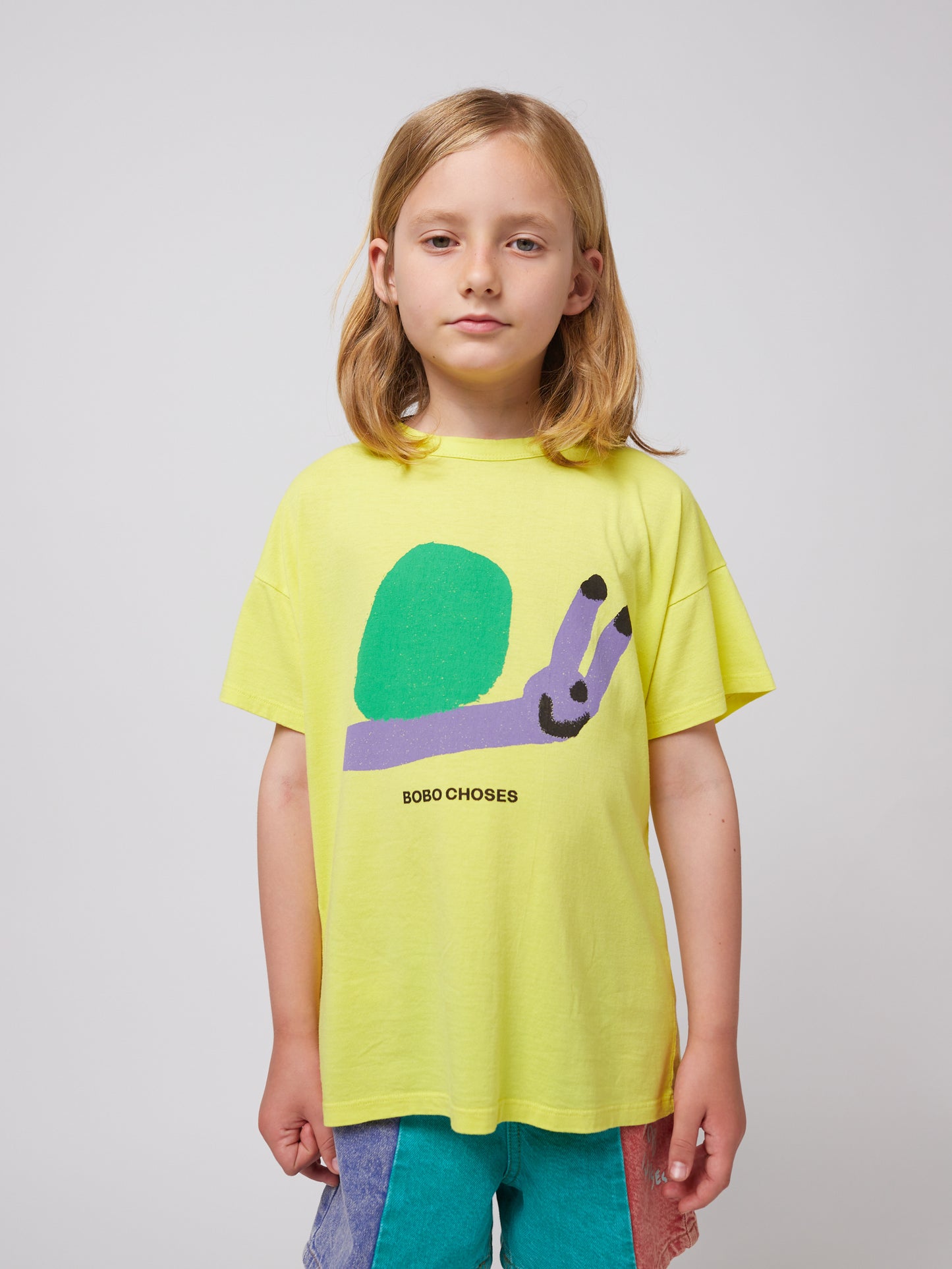 A child wearing the Bobo Choses Funny Snail T-Shirt in bright yellow. The T-shirt features a playful illustration of a smiling snail with a green shell and a lavender body, along with "BOBO CHOSES" printed in black below the design. The child is also wearing multicoloured shorts.