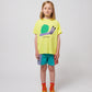 A child wearing the Bobo Choses Funny Snail T-Shirt in bright yellow. The T-shirt features a playful illustration of a smiling snail with a green shell and a lavender body, with "BOBO CHOSES" printed below. The child pairs it with multicoloured shorts, orange socks with sunflower patterns, and translucent orange jelly sandals.