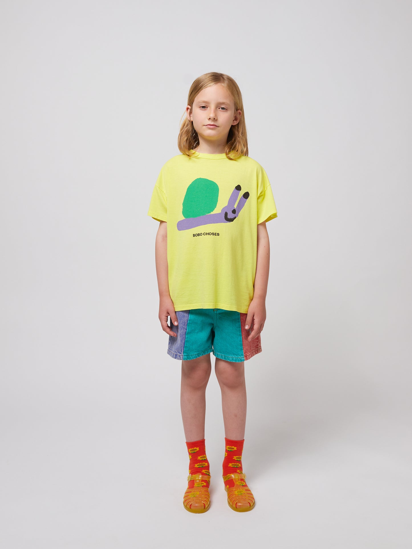 A child wearing the Bobo Choses Funny Snail T-Shirt in bright yellow. The T-shirt features a playful illustration of a smiling snail with a green shell and a lavender body, with "BOBO CHOSES" printed below. The child pairs it with multicoloured shorts, orange socks with sunflower patterns, and translucent orange jelly sandals.