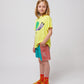 A child wearing the Bobo Choses Funny Snail T-Shirt in bright yellow, featuring a playful illustration of a smiling snail with a green shell and lavender body. The child is styled in multicoloured shorts with a red and blue patchwork design, orange sunflower-patterned socks, and translucent orange jelly sandals. The side view showcases the relaxed fit of the T-shirt and the vibrant outfit combination.