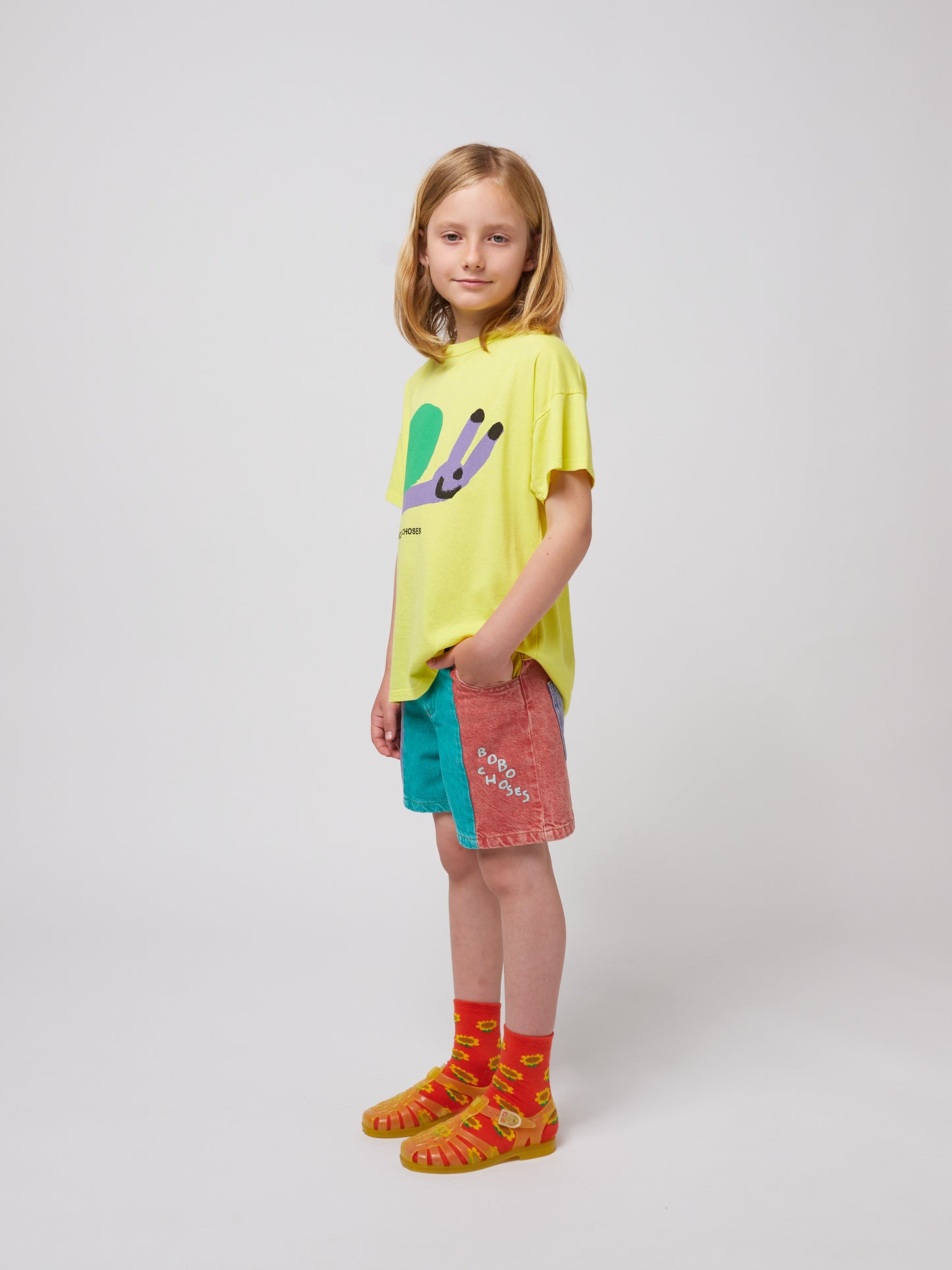 A child wearing the Bobo Choses Funny Snail T-Shirt in bright yellow, featuring a playful illustration of a smiling snail with a green shell and lavender body. The child is styled in multicoloured shorts with a red and blue patchwork design, orange sunflower-patterned socks, and translucent orange jelly sandals. The side view showcases the relaxed fit of the T-shirt and the vibrant outfit combination.