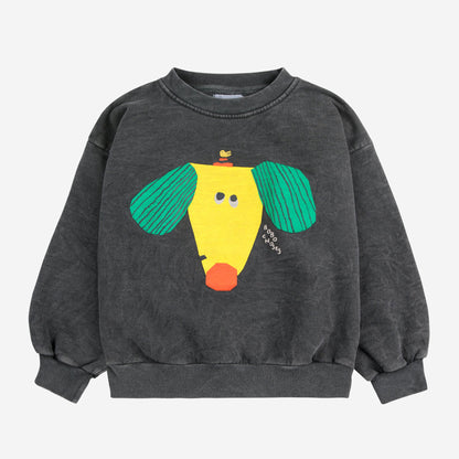 A charcoal grey children's sweatshirt featuring a playful, abstract graphic of a yellow dog with green ears and an orange snout. A small bird is perched on its head, and the "Bobo Choses" logo is printed beside the design. The sweatshirt has a relaxed fit, ribbed cuffs, and a ribbed hem.