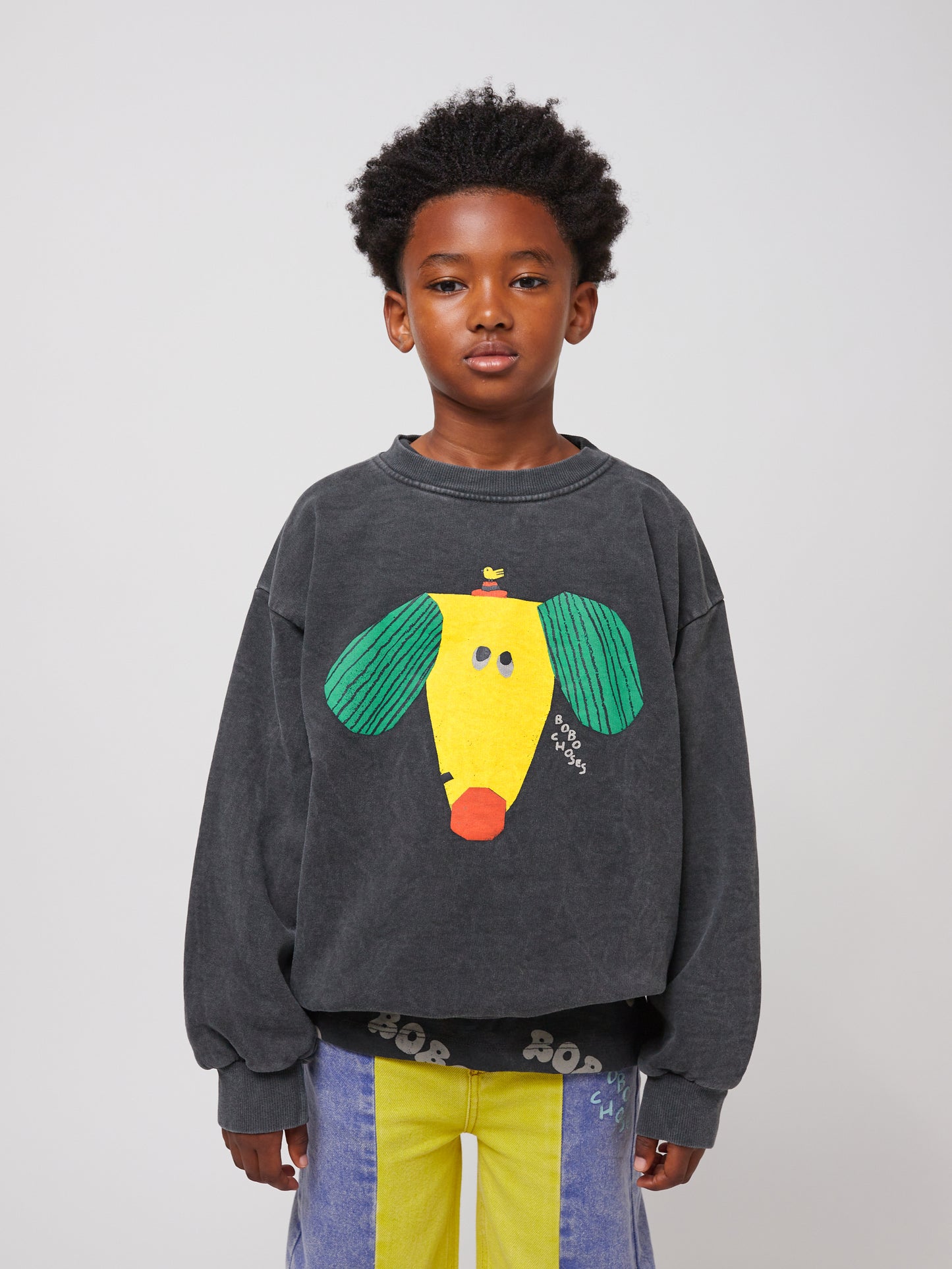 A child with short, curly hair wears a charcoal grey oversized sweatshirt featuring a playful, abstract dog illustration in bright yellow, green, and red, with "BOBO CHOSES" written on it. The sweatshirt has a relaxed fit with dropped shoulders and ribbed cuffs. The child also wears wide-leg, colour-blocked denim jeans in yellow, blue, and red.