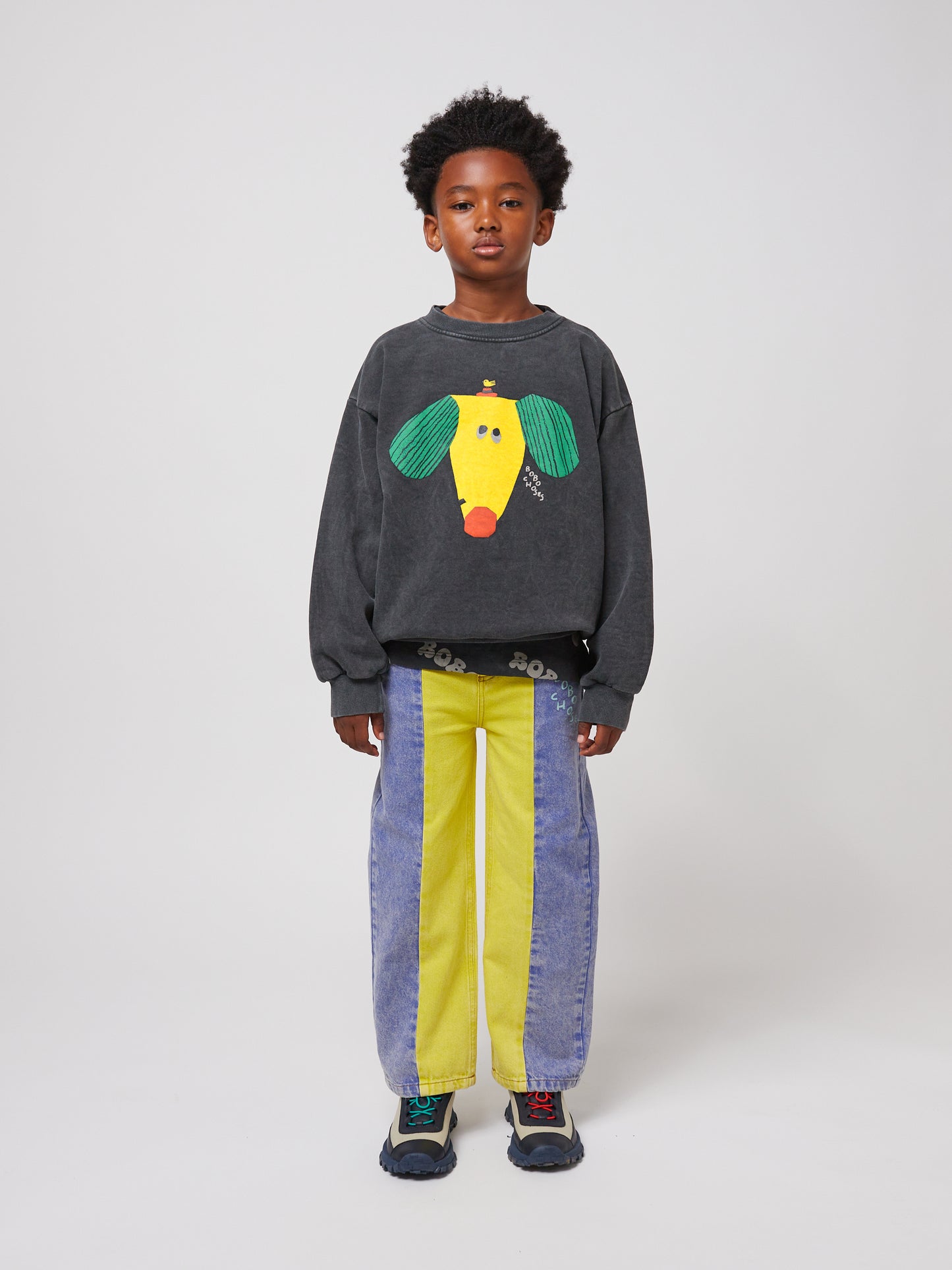 Full-body shot of the same child standing against a plain white background. They wear a dark grey sweatshirt with a colourful dog illustration, paired with two-tone denim jeans and chunky multicoloured trainers.