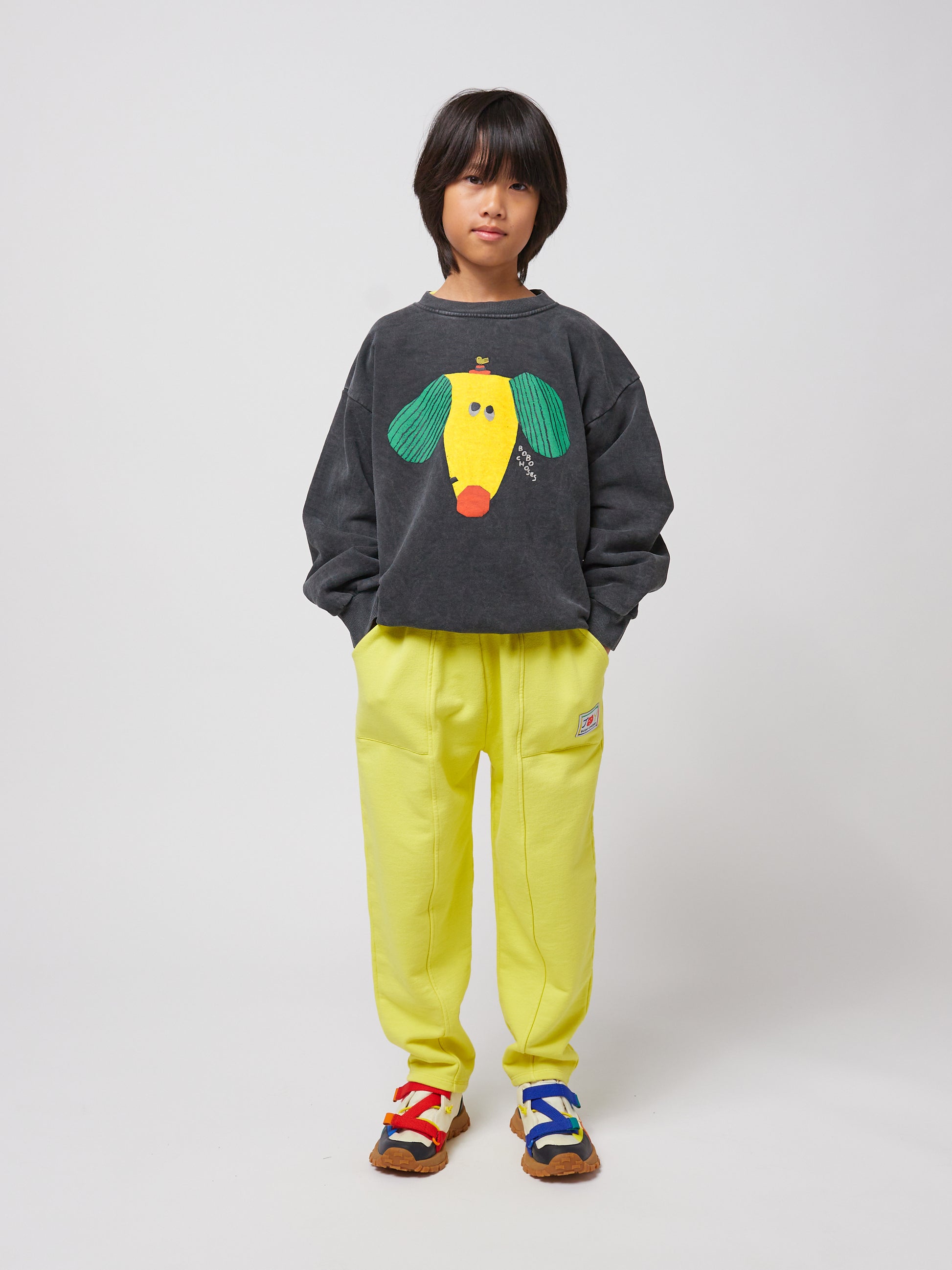 Another child, with medium-length dark hair, wears the same dark grey sweatshirt but pairs it with vibrant yellow sweatpants and multicoloured trainers with red, blue, and white straps.