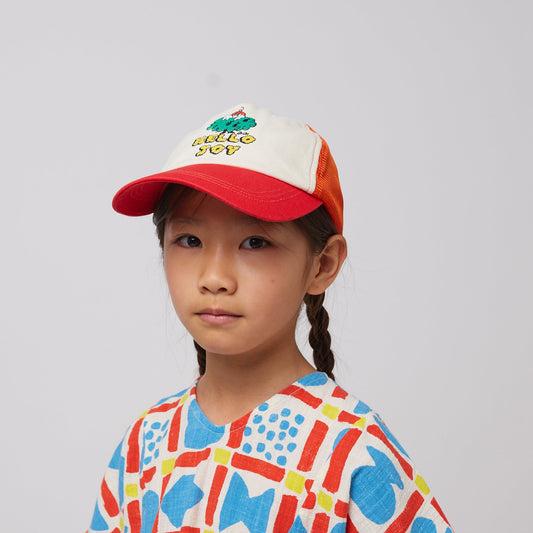 A child wearing the Bobo Choses "Hello Joy" mesh cap, featuring a cream front panel with an embroidered tree and "HELLO JOY" text in yellow. The cap has a red brim and orange mesh back. The child is also wearing a geometric-print top in red, blue, and cream.
