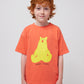 The child is shown from the waist up, wearing the coral T-shirt with the yellow bear print. Their red hair and freckles are visible, giving a cheerful look to the image.