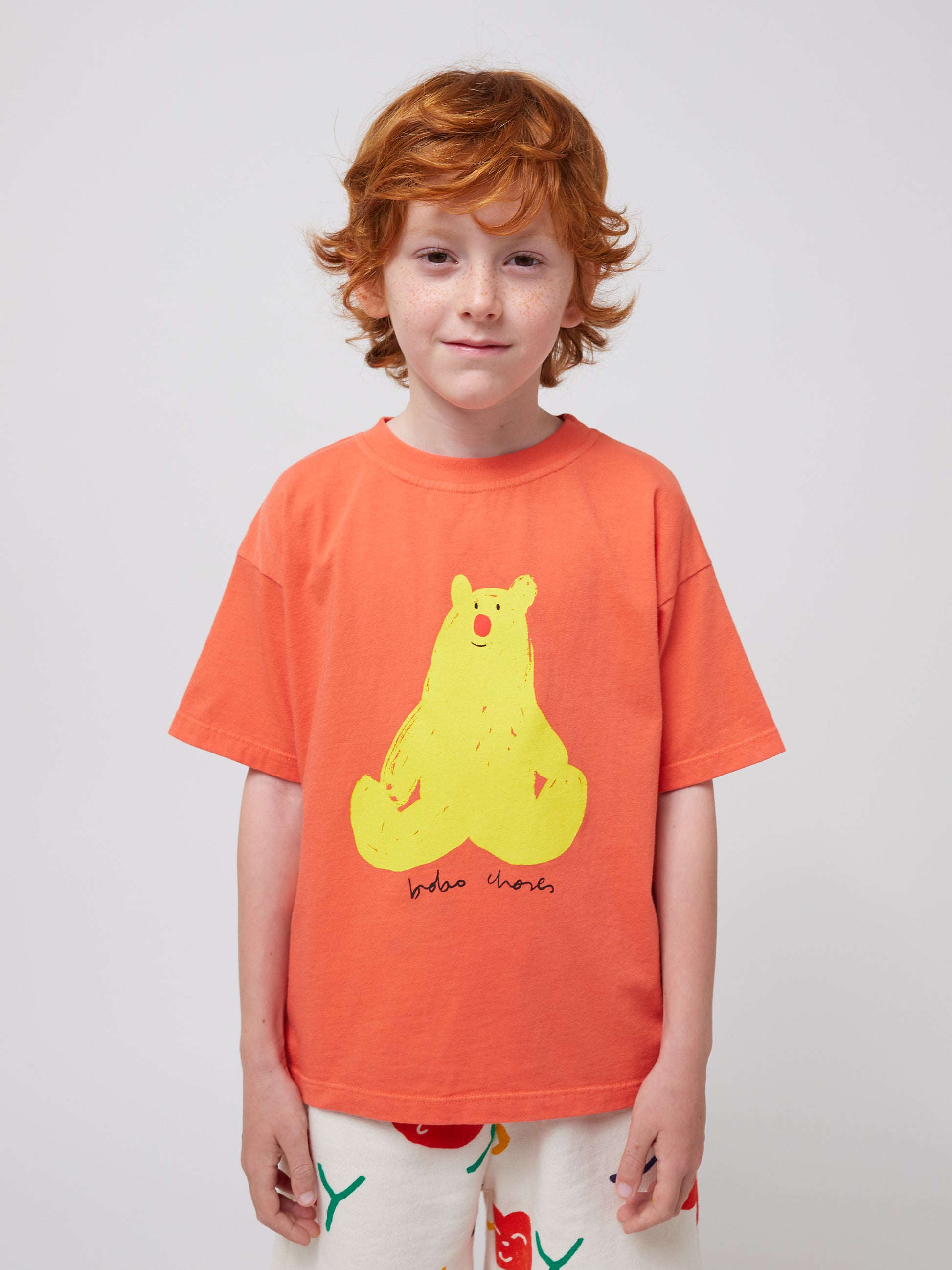 The child is shown from the waist up, wearing the coral T-shirt with the yellow bear print. Their red hair and freckles are visible, giving a cheerful look to the image.