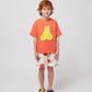 The child stands front-facing in the oversized coral T-shirt with a yellow bear print, paired with white shorts featuring a red cherry design. Their outfit is completed with multicoloured sandals.