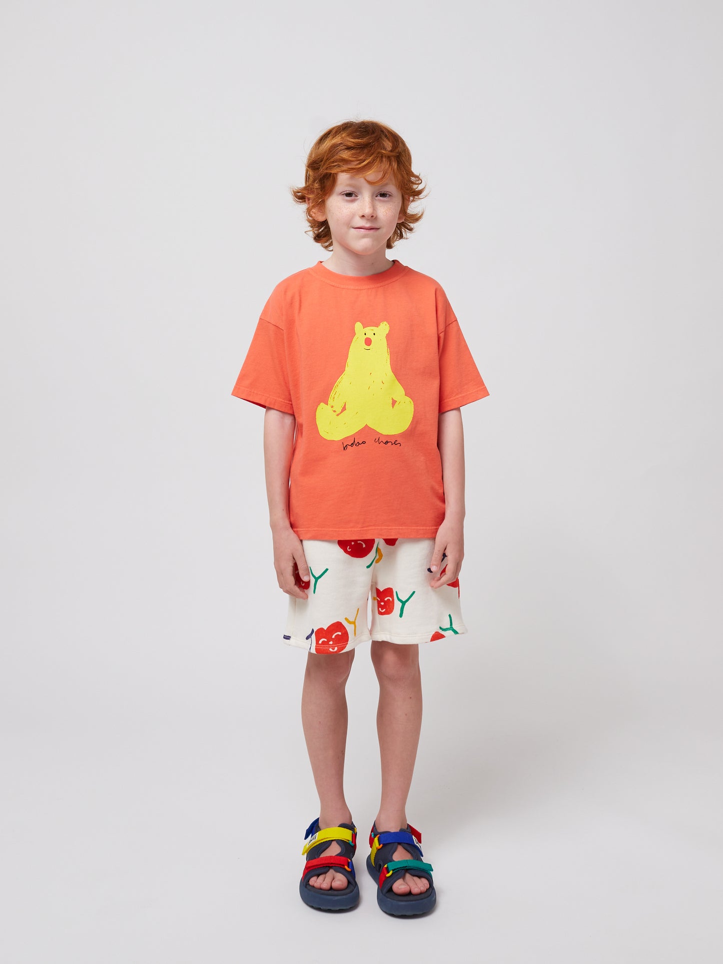 The child stands front-facing in the oversized coral T-shirt with a yellow bear print, paired with white shorts featuring a red cherry design. Their outfit is completed with multicoloured sandals.