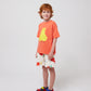 A child with curly red hair wears the coral Bobo Choses T-shirt with white shorts featuring a cherry print. They are also wearing multicoloured sporty sandals.
